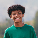 A teenage boy with curly hair wearing a green t-shirt, smiling